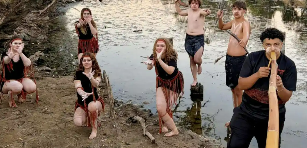 Image features seven aboriginal youth outside by a creek. There are four girls striking dance poses on the right side of the image, they have traditional face paint and woven dance costumes. On the right side of the image there is one boy playing a didgerydoo and behind him are two more boys balanced on logs in the edge of the water holding traditional hunting weapons, all three boys also have traditional face paint.