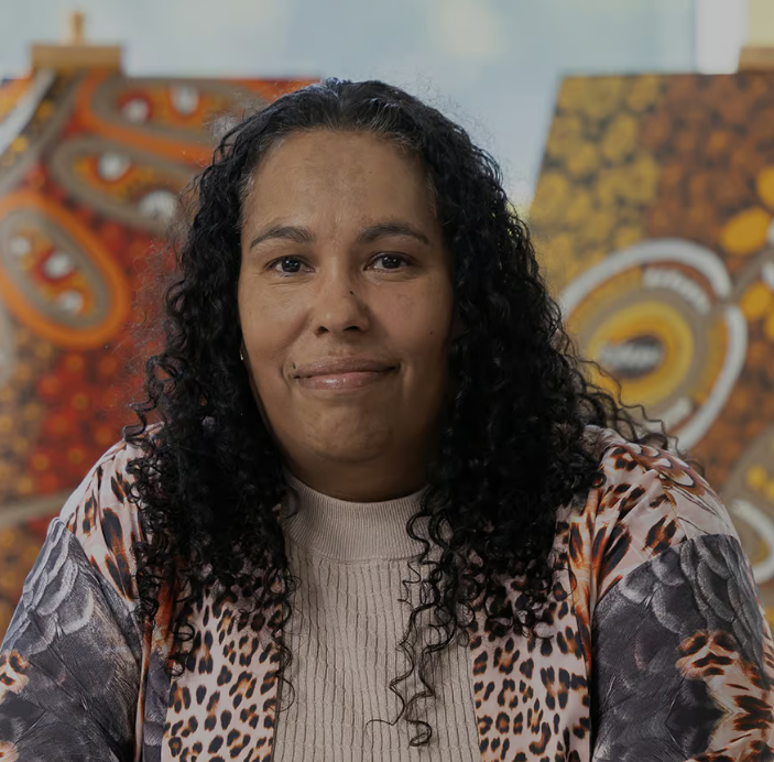 The image shows the head and shoulders of Noongar woman Fiona Reidy, she is middle aged and has black curly hair down past her shoulders, she is smiling at the camera. In the background are two of her artworks in dot painting style in yellow, oragne, and brown colours.