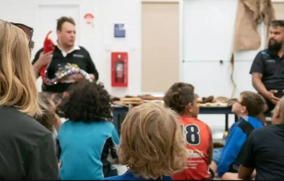 Image shows two men in shirts with cultural designs telling a story to a group of youth and an adult, one of the men holds a red toy snake