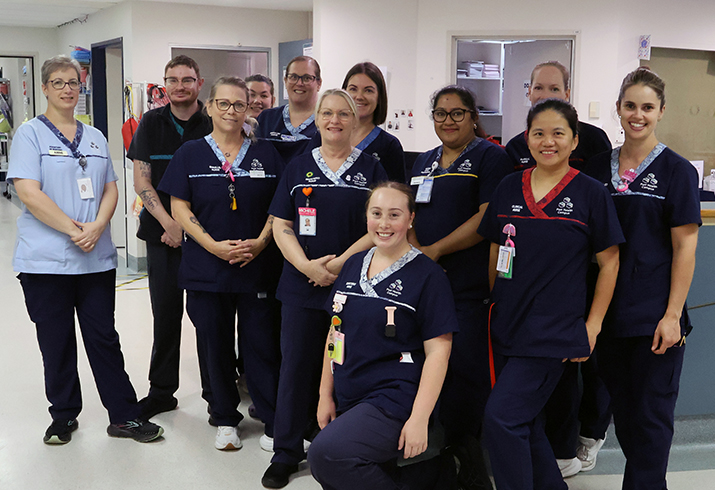 image contains 12 healthcare staff in a group inside a hospital, 11 staff are dressed in in dark blue hospital uniforms and one manager in a light blue uniform stood on the left of the group as we look at them